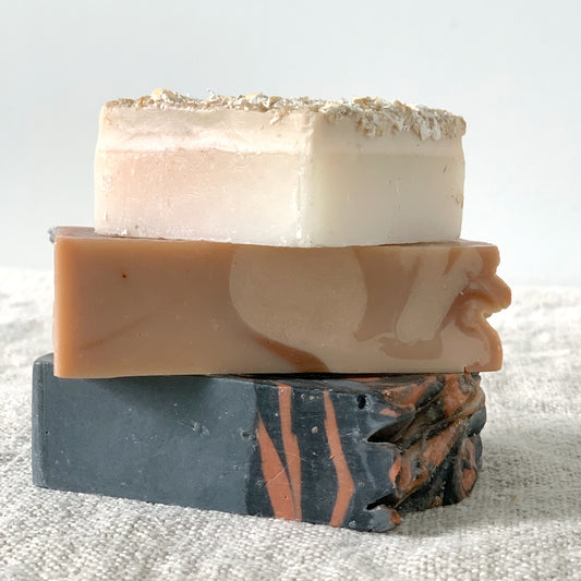 Stack of handmade soaps on a linen towel. Top soap is square and cream colored with oat texture on top. Middle soap is rectangular and rose - white swirled in color. The bottom soap is also rectangular, is a dark gray and clay swirled colored.  