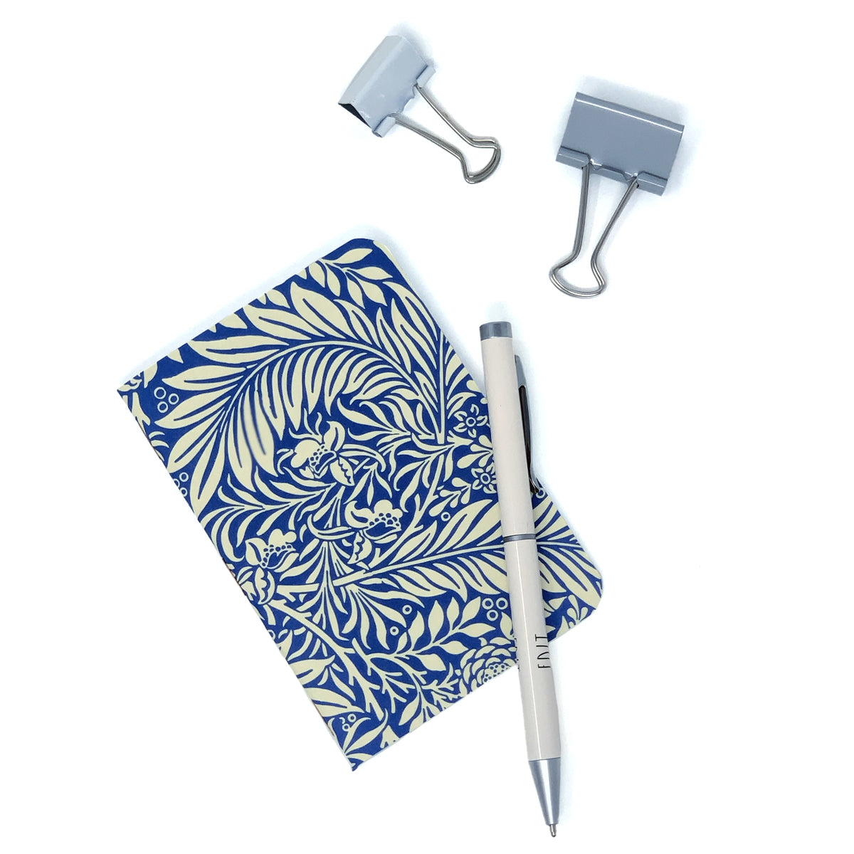 Pocket notebook with William Morris blue and off white floral pattern on cover. 2 binder clips and white pen sit next to the notebook on a white background.