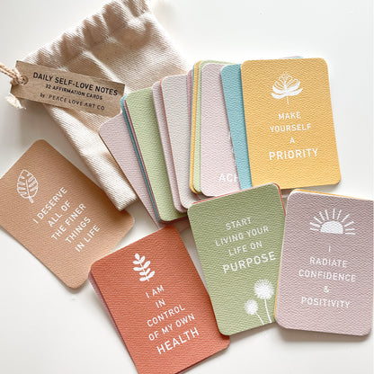 Colorful deck of affirmation cards, each printed with a positive message and nature-inspired illustration. Scattered cards on top of a handmade cotton pouch against a white background.